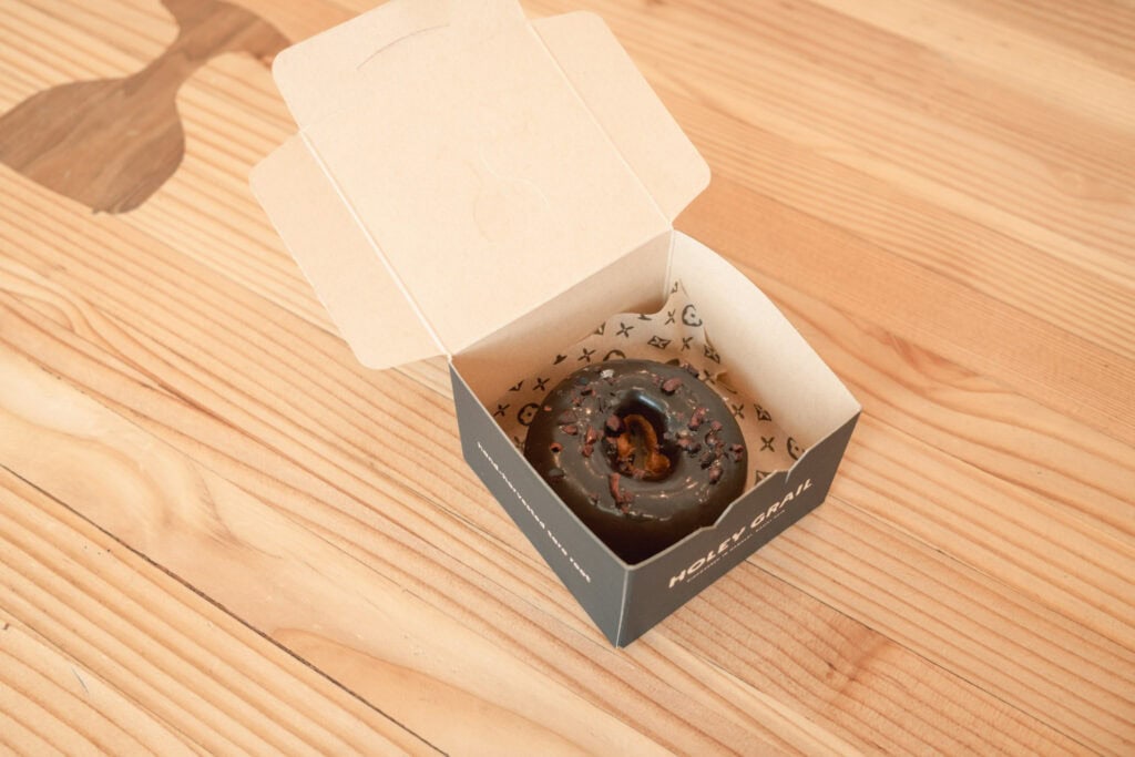 Holey Grail Donuts 的巧克力甜甜圈裝在紙盒中,放在木桌上。