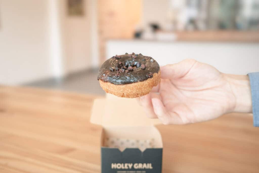 手拿一顆 Holey Grail Donuts 的巧克力脆片甜甜圈,底下是品牌紙盒。