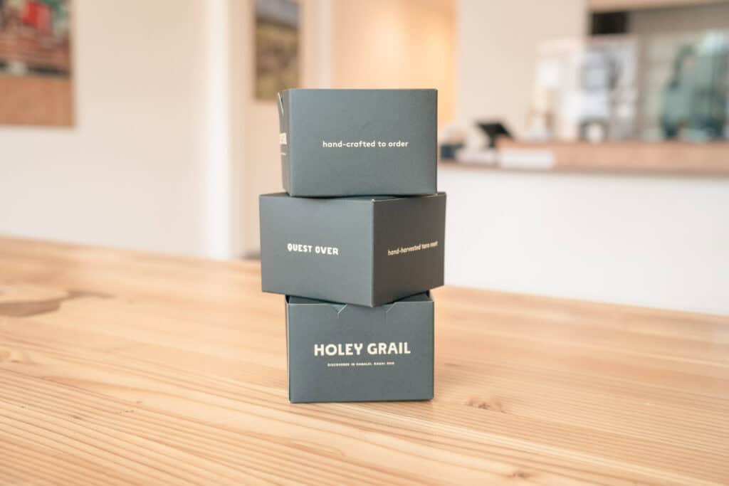 三個印有 Holey Grail Donuts 標語的甜甜圈紙盒堆疊在木質桌面上